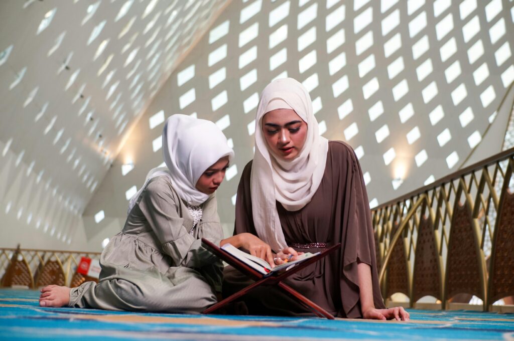 A mother and daughter wearing hijabs reading the Quran in a beautifully lit mosque interior.
